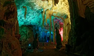 Khao Pueng cave chamber illuminated with vibrant artificial lights in green, blue, red, and yellow, showcasing stalactites, stalagmites, and textured rock walls.