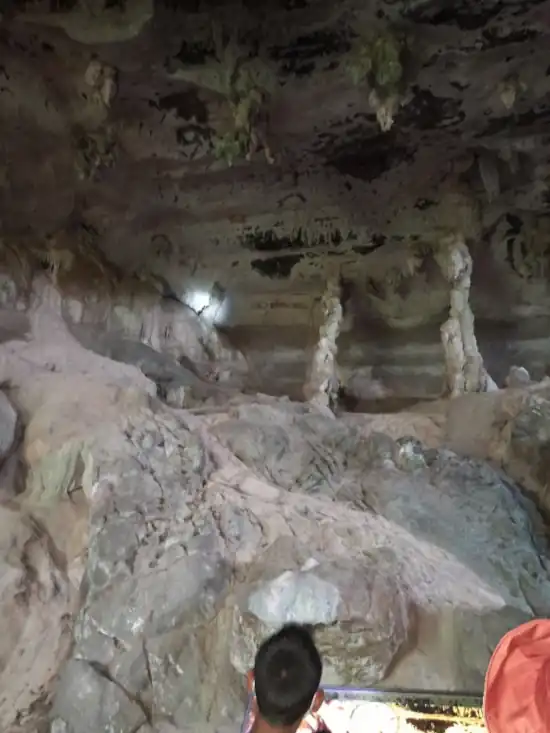A cave chamber with towering stalactites and stalagmites, illuminated to highlight textures, with a person standing below to show the scale of the formations.