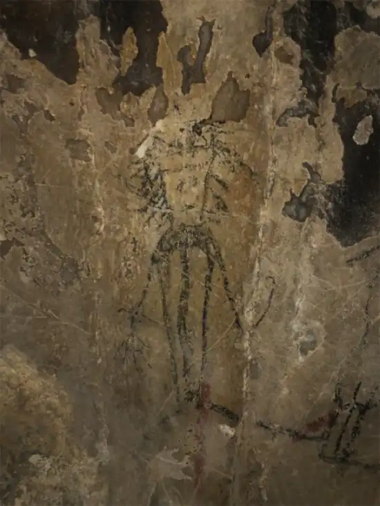 Rock art showing a humanoid figure with extended arms and elongated legs, drawn in black pigment on a weathered stone wall.