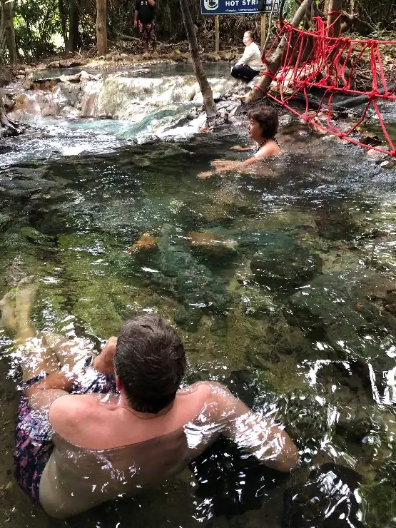 A natural hot spring in a forest setting, where people relax in the clear shallow water, surrounded by rocks and tropical vegetation.