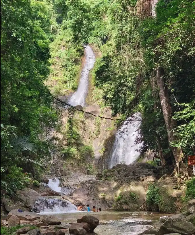 A multi‑level waterfall flowing through a rocky cliff into a shallow pool, with people wading at the base amid lush tropical forest.