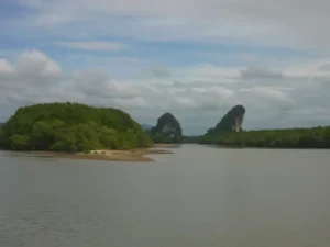 The Krabi river estuary bordered by dense mangrove forest, with two dramatic limestone karst formations rising in the background under a partly cloudy sky.
