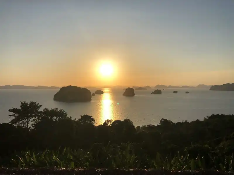 A serene sunset view over calm water, with golden reflections, silhouetted rocky islands, and lush vegetation in the foreground.