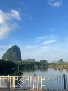 Scenic view of calm water reflecting lush greenery and towering limestone hills, with a white railing in the foreground under a sunny sky.