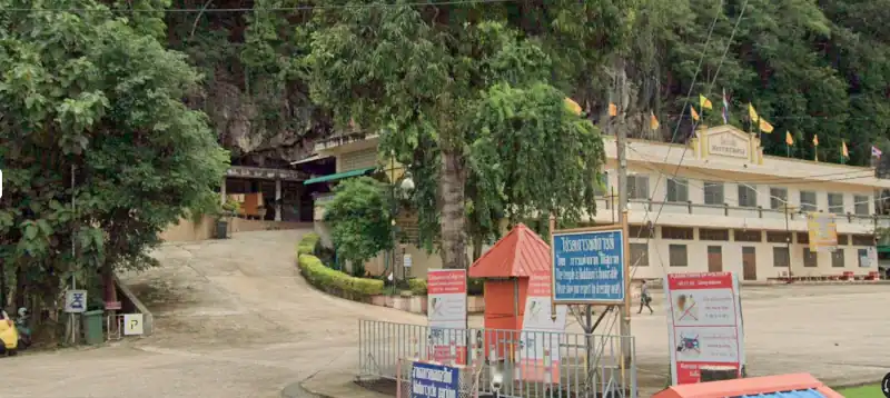 Entrance area with buildings against a rocky hillside, flags on display, information boards, and a scooter parked near signage.