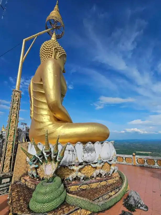 Large golden Buddha seated on ornate pedestal with serpent figures, decorative umbrella above, and panoramic landscape backdrop.